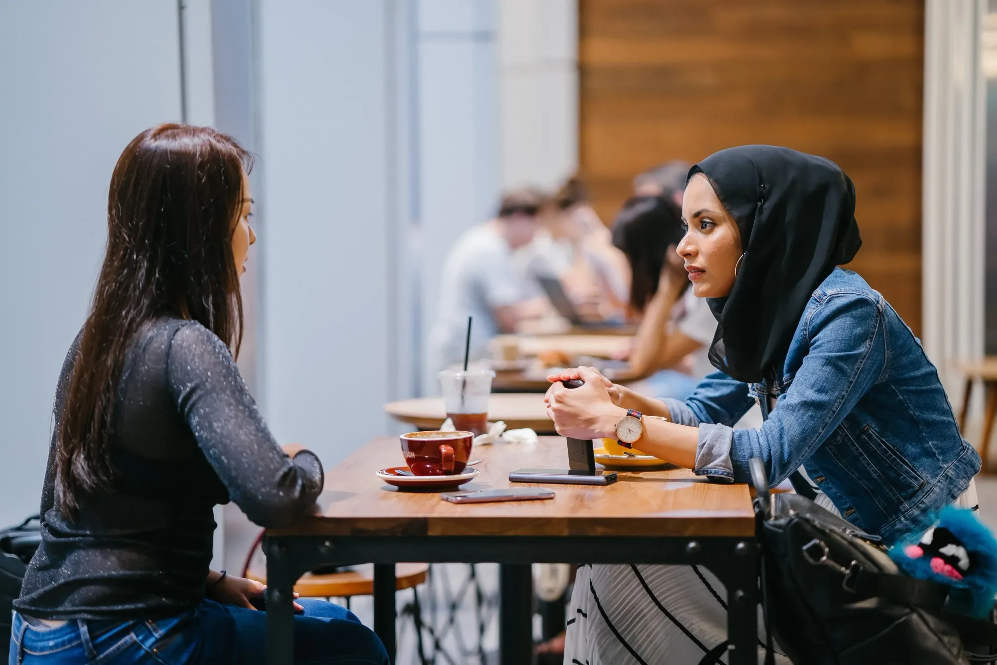 Two educators in conversation over coffee at a café table, one wearing a hijab